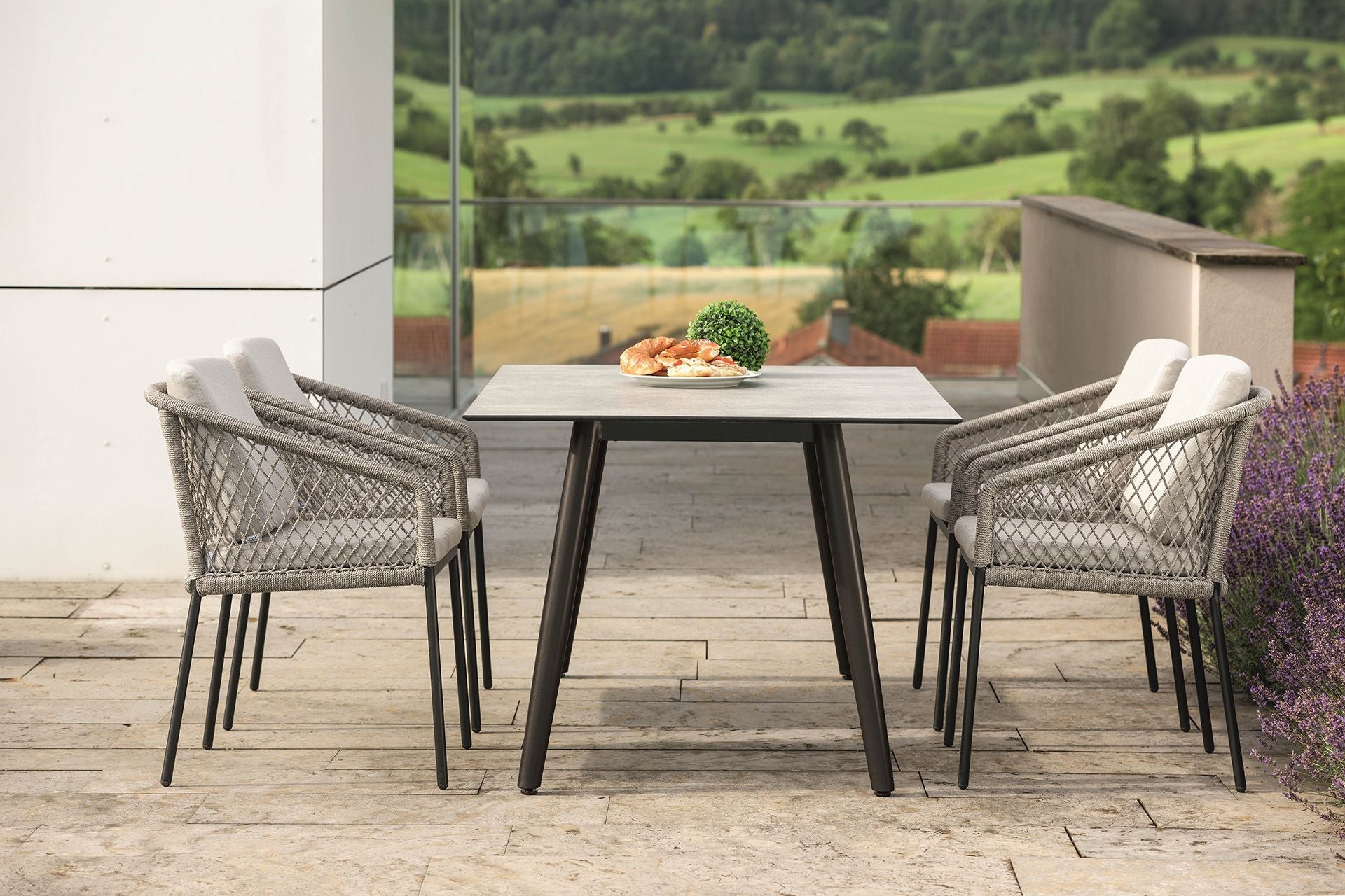 Moderner Outdoor-Esstisch mit vier geflochtenen Sesseln und Polstern auf Terrasse mit Ausblick ins grüne Tal, gedeckt mit Gebäck und Deko.