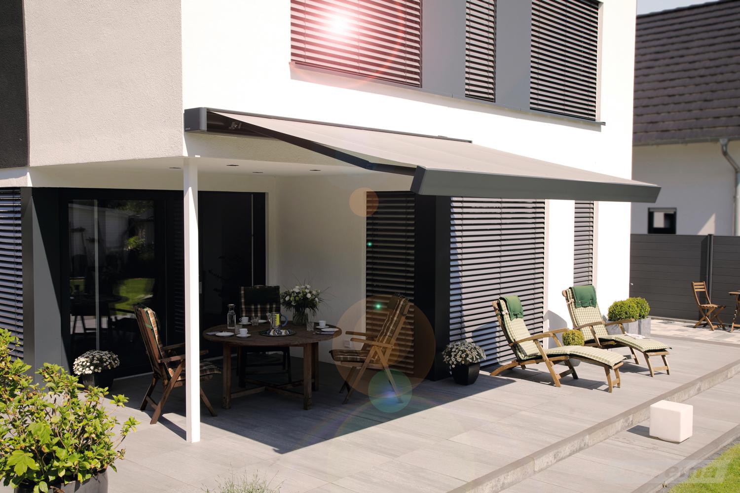 Moderne Kassettenmarkise in Grau spendet Schatten über einer Terrasse mit Esstisch und zwei Holzliegen an einem weißen Einfamilienhaus.