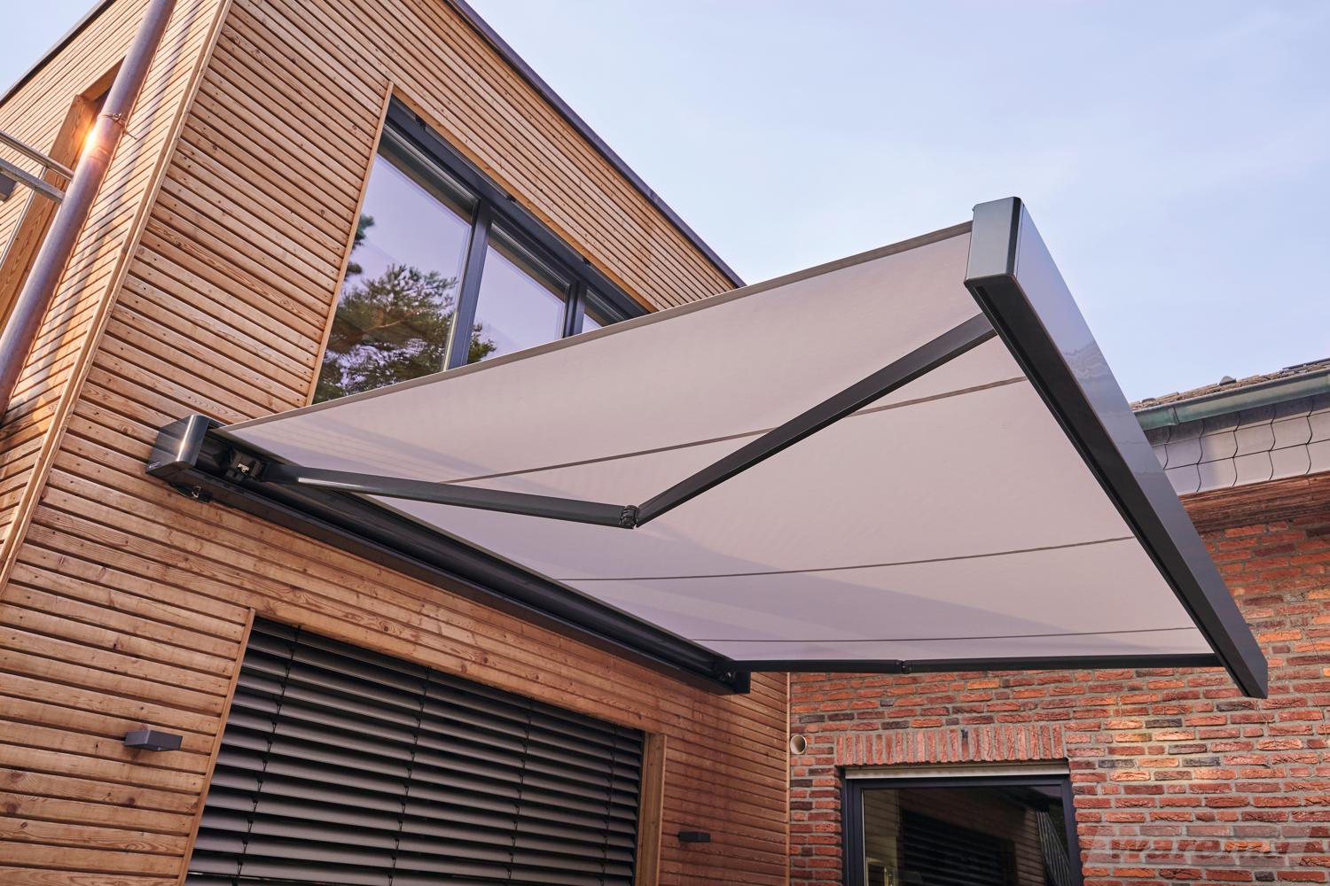 Gelenkarmmarkise in eleganter, neutraler Farbe an einer Holzfassade montiert, bietet Schatten auf einer modernen Terrasse.