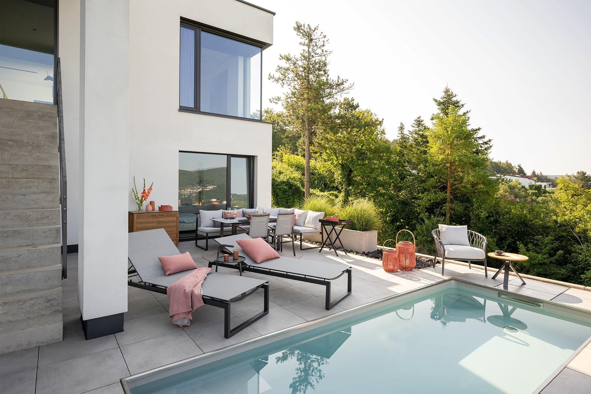 Moderne Terrasse mit Pool, Sonnenliegen und stilvollen Outdoor-Möbeln von BOKORA; umgeben von grüner Natur und mit Blick ins Tal.