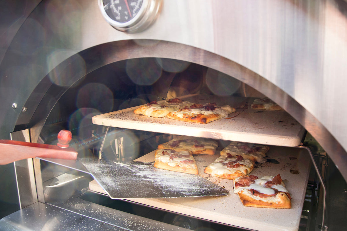 Pizza mit Schinken und Käse auf zwei heißen Pizzasteinen in einem Edelstahl-Pizzaofen, Schieber aus Holz und Metall im Einsatz.