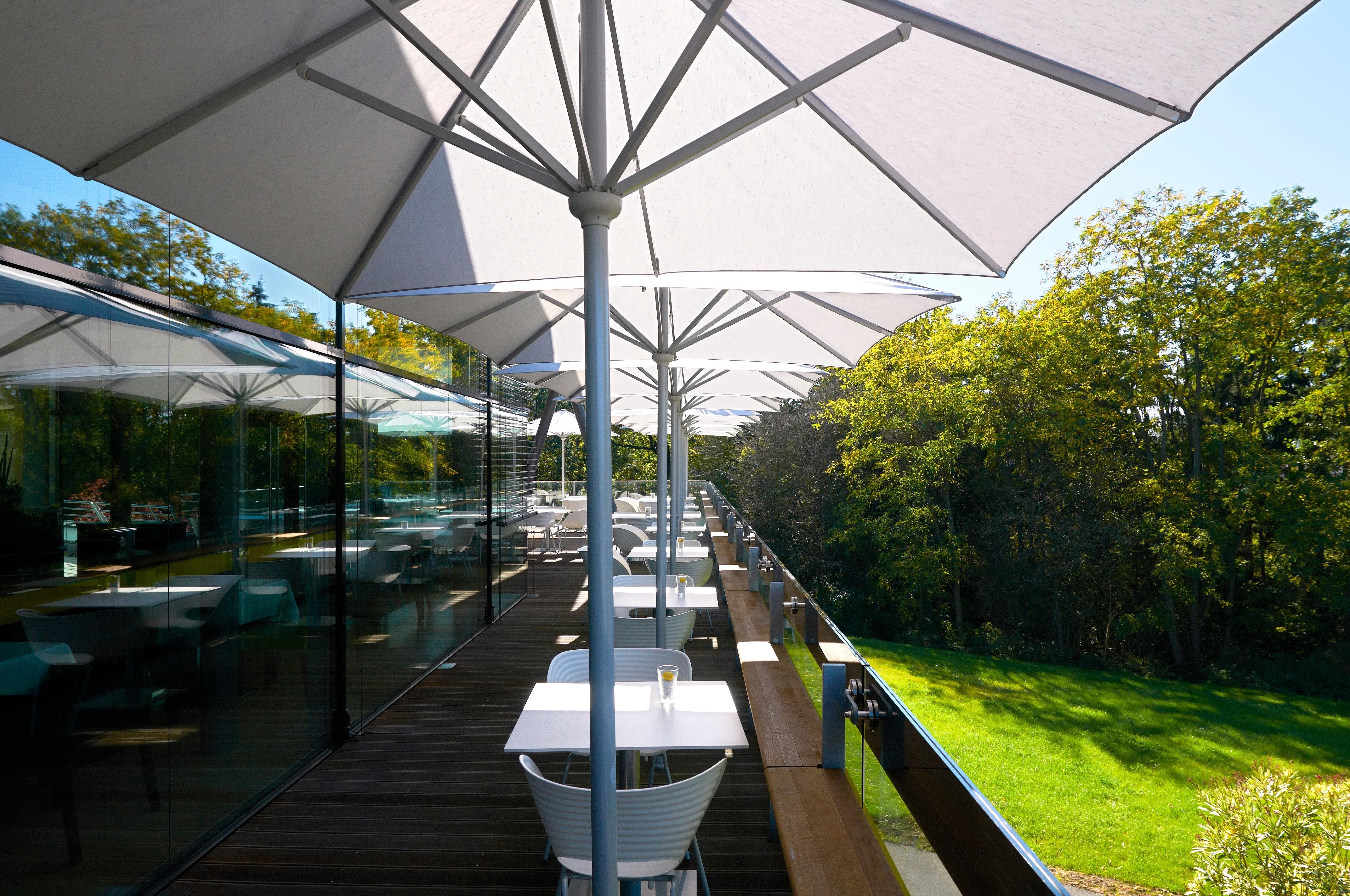 Reihe großer weißer Mittelmast-Sonnenschirme auf einer modernen Restaurantterrasse mit Glasfassade und Blick ins Grüne.