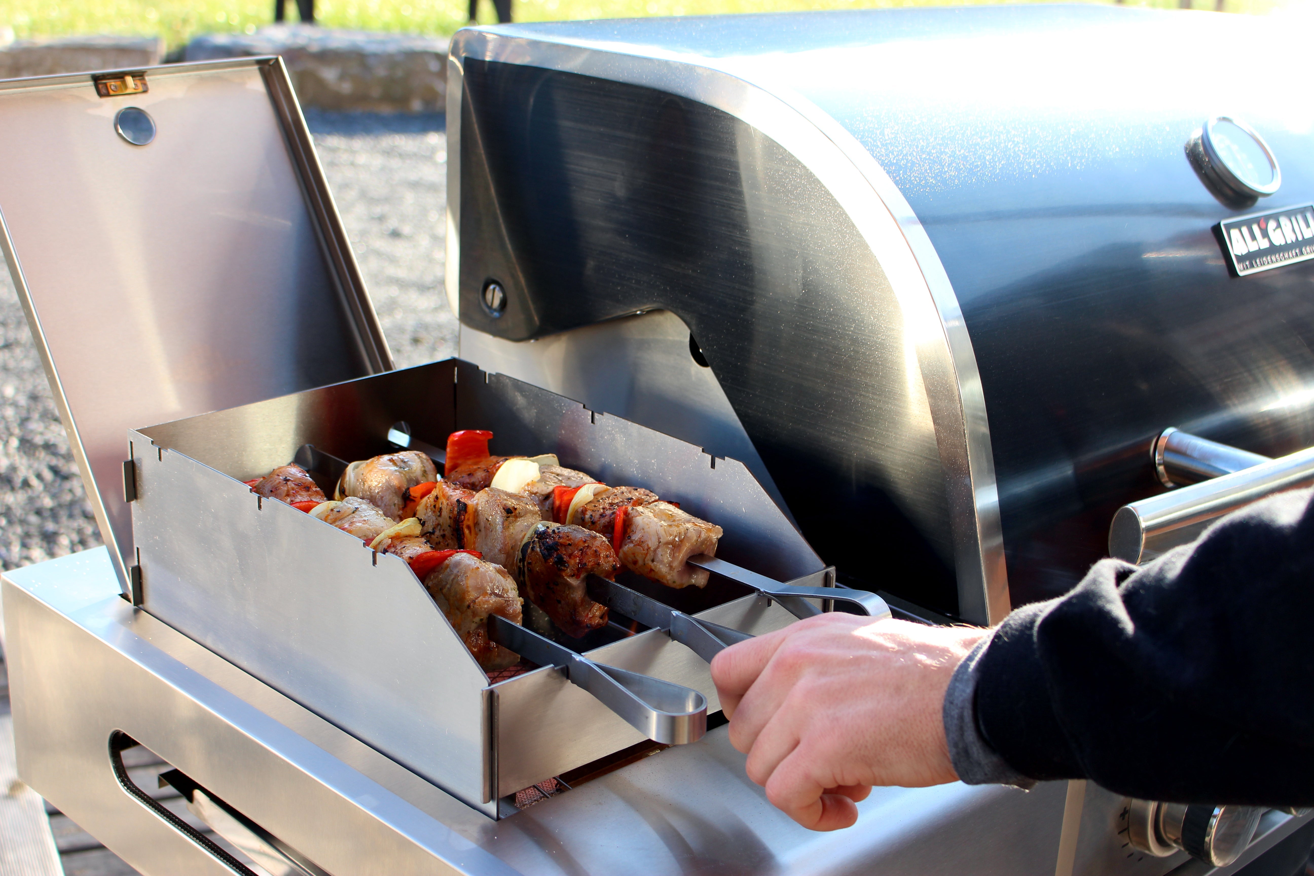 Nahaufnahme eines ALLGRILL Gasgrills mit Grillspießen, auf denen Fleisch und Gemüse gegrillt werden; Hand dreht die Spieße.