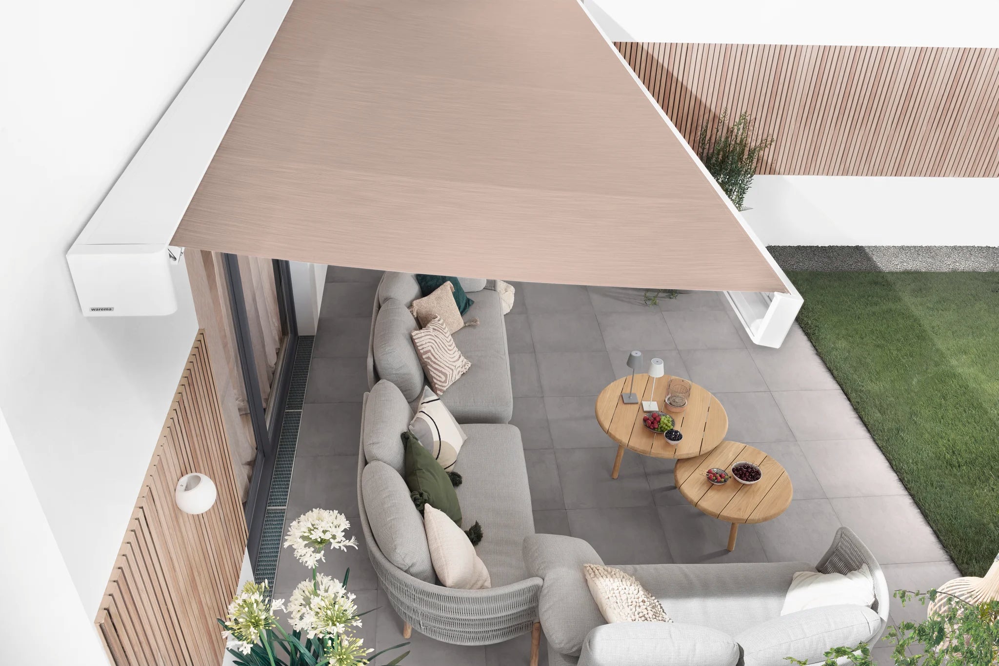 Moderne Markise in hellem Beige über einer gemütlichen Outdoor-Lounge mit Sofas und Holztischen auf einer Terrasse mit Gartenanschluss.