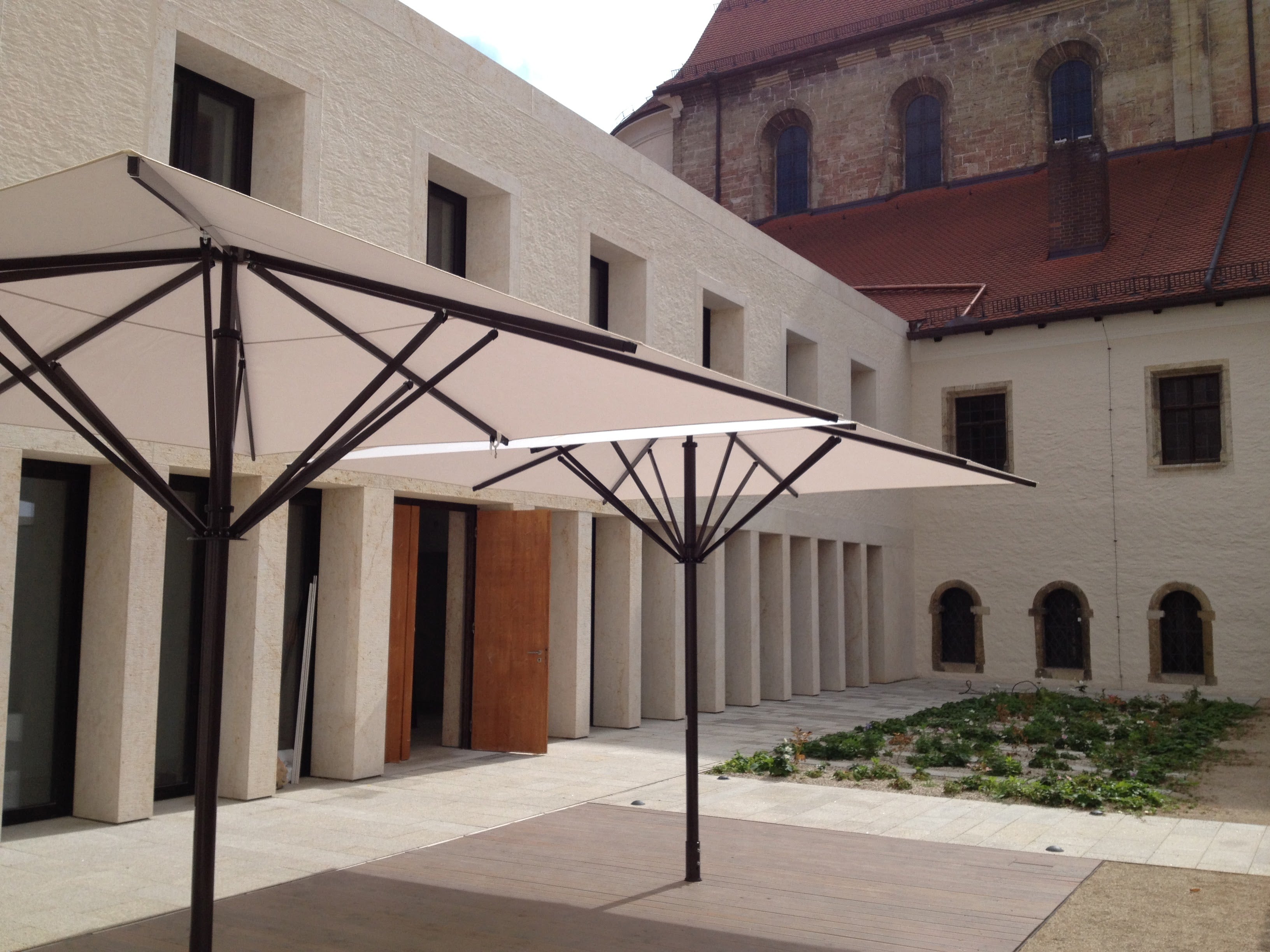 Modernes Gebäude neben einer alten Kirche mit großen Gastro-Sonnenschrimen mit hellem Stoff und schwarzem Mast.
